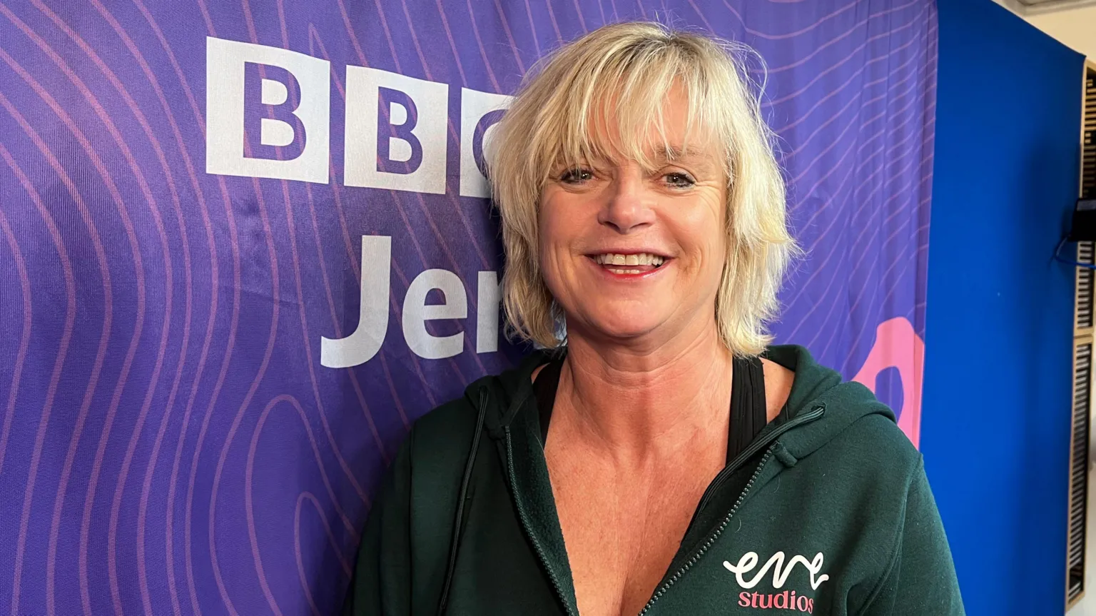 Image shows Trudi Roscouet wearing a black hoody, with 'eve studios' branding, smiling with short blond hair, beside a BBC Jersey purple backdrop.