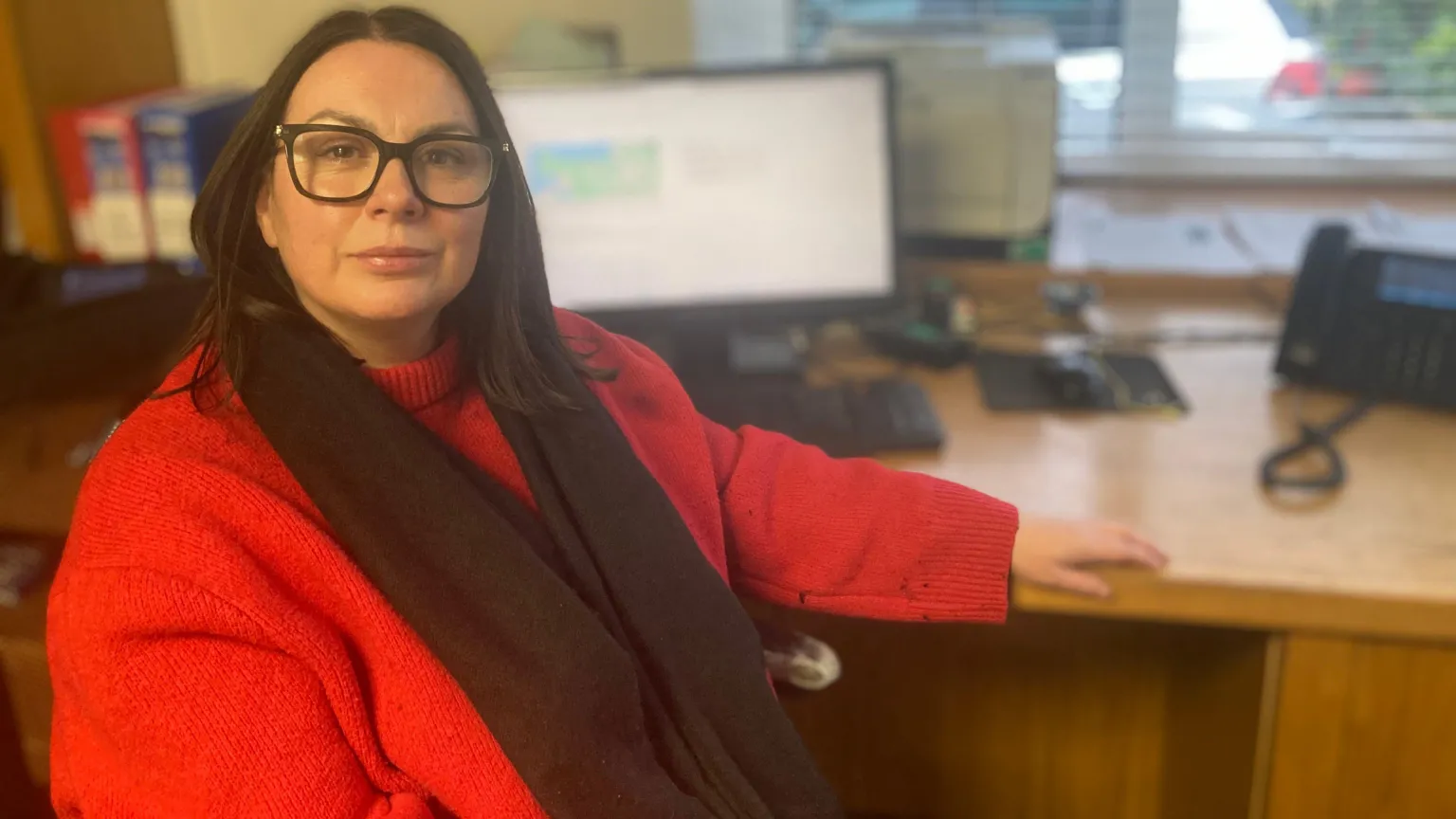 Bernie is wearing a red jumper and a maroon scarf. She's sitting at a wooden table. One of her hands is resting on the table. A computer and landline phone are on the table. Some files are on a shelf.