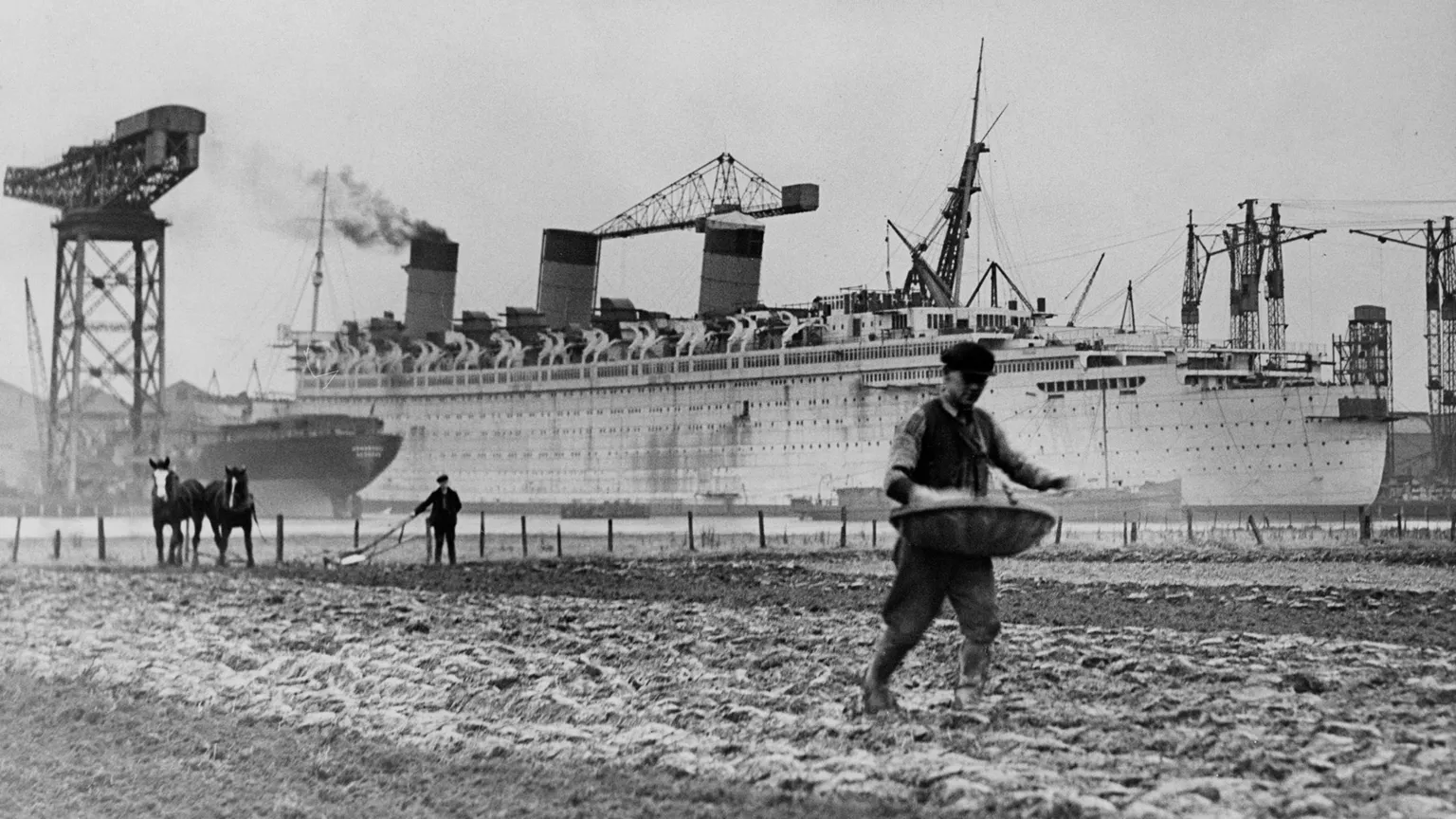  A black and white image of a large ocean liner with a large crane in the left of the shot and people sowing fields in the foreground
