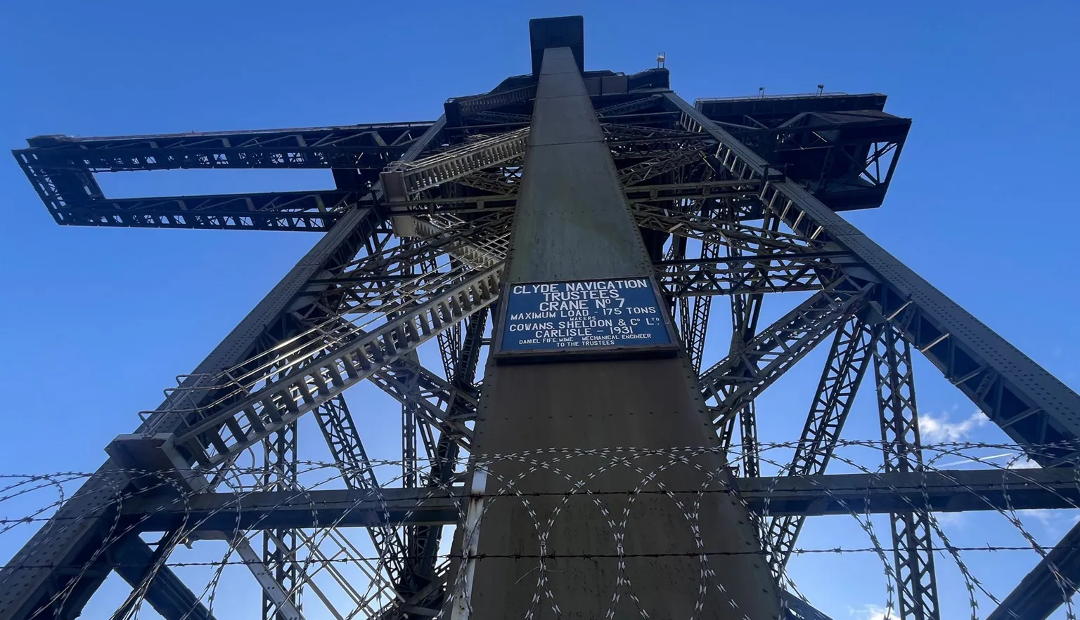 A large latticed steel crane photographed looking up, with razor wire and the name of the crane on the elevator