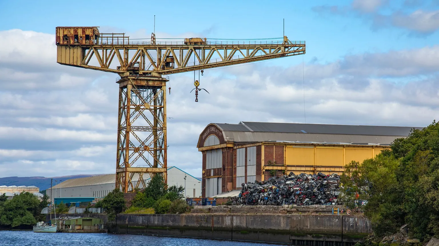  A yellow steel latticed crane beside a river. Beside it is a modernist style industrial building
