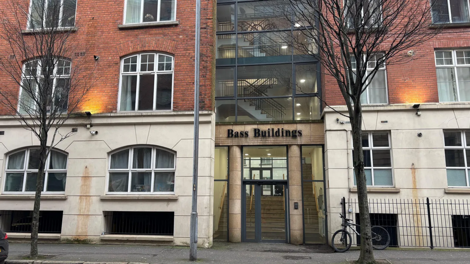 A photo of the entrance to the Bass Buildings apartment complex in Belfast city centre. The six storey residential building is a mix of red brick and grey or pale render or blocks. It has large arched windows and there are two stone or concrete columns on either side of the glass front doors. A set of grey steps are visible through the doors. There is a a large column of windows above the entrance, through which can be seen a tiered staircase running up the middle of the building.