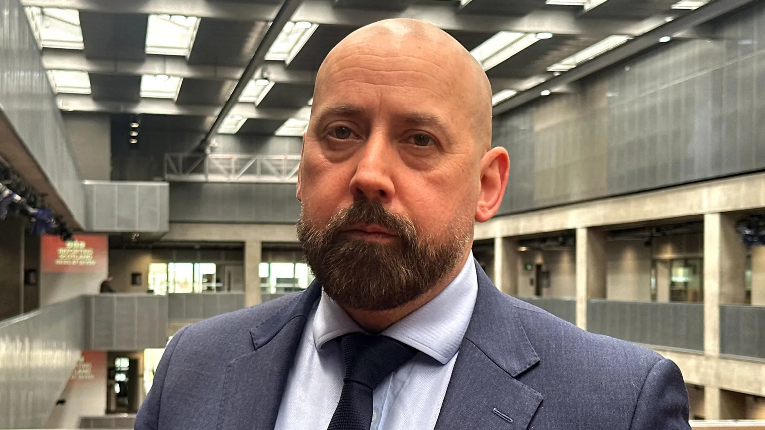A man with a beard wearing a dark suit and a tie, looking directly at the camera