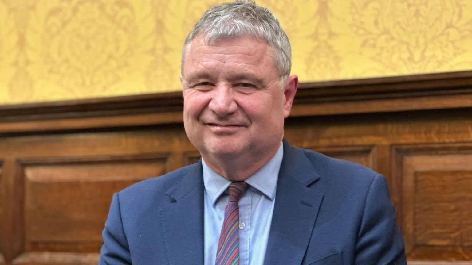 Chris Thomas, who has grey hair and is wearing a blue suit with a stripped tie, in the Tynwald chamber. There is wood panelling and yellow patterned wallpaper behind him.