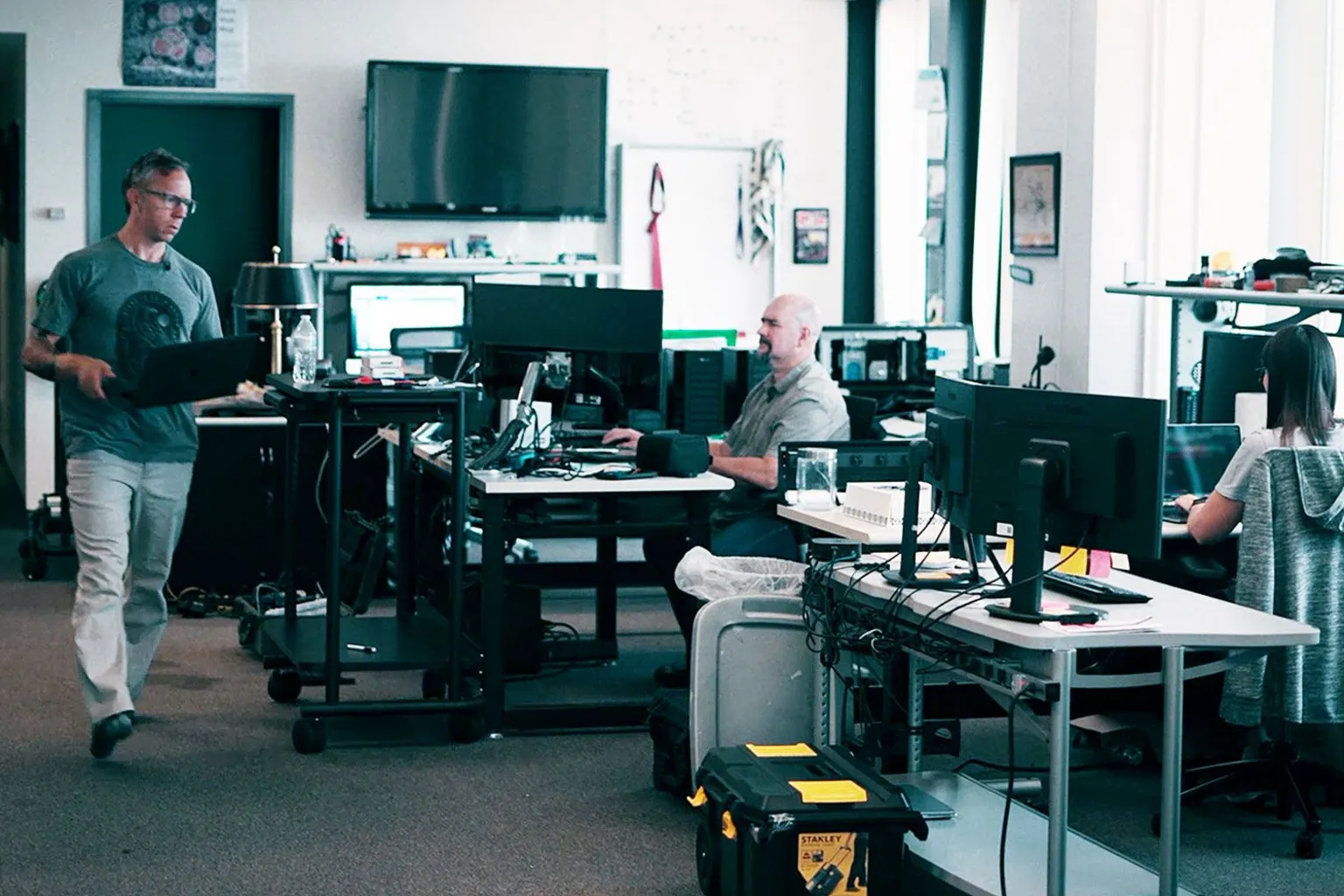 Greg Squire and colleagues including Pete Manning in their office. Greg is wearing a green t-shirt and light trousers and is walking across a room crowded with computer screens, holding a laptop. 