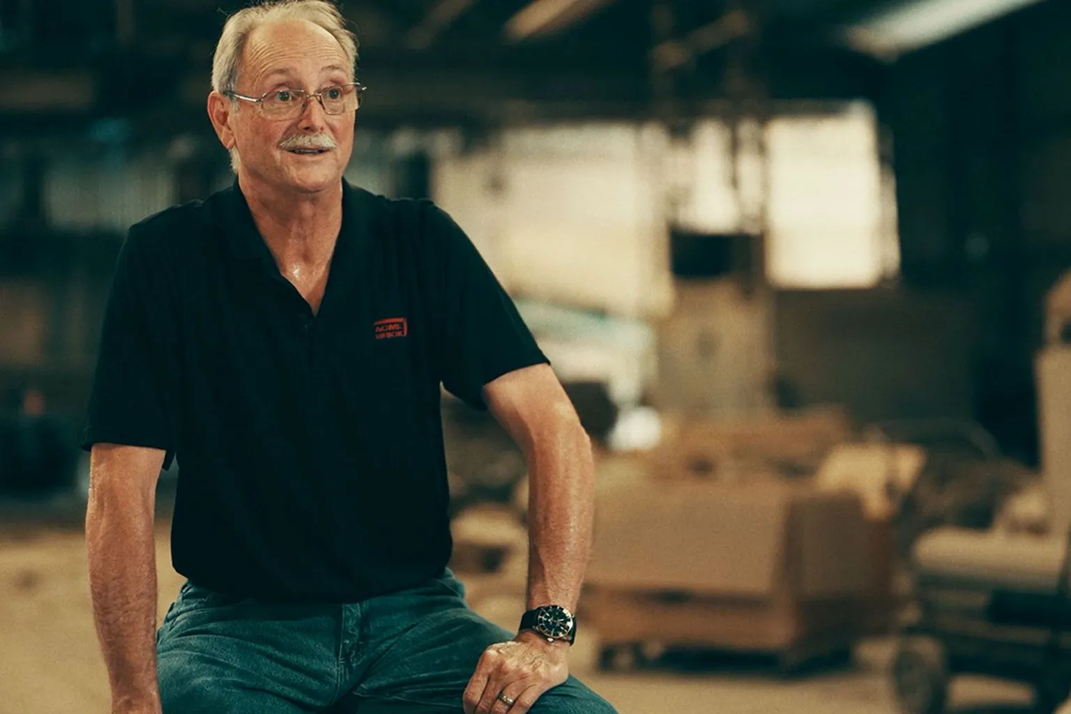 John Harp has short grey hair and a grey moustache. He is wearing a black short-sleeved shirt and green trousers, and is sitting in his brick factory, open boxes open behind him. 
