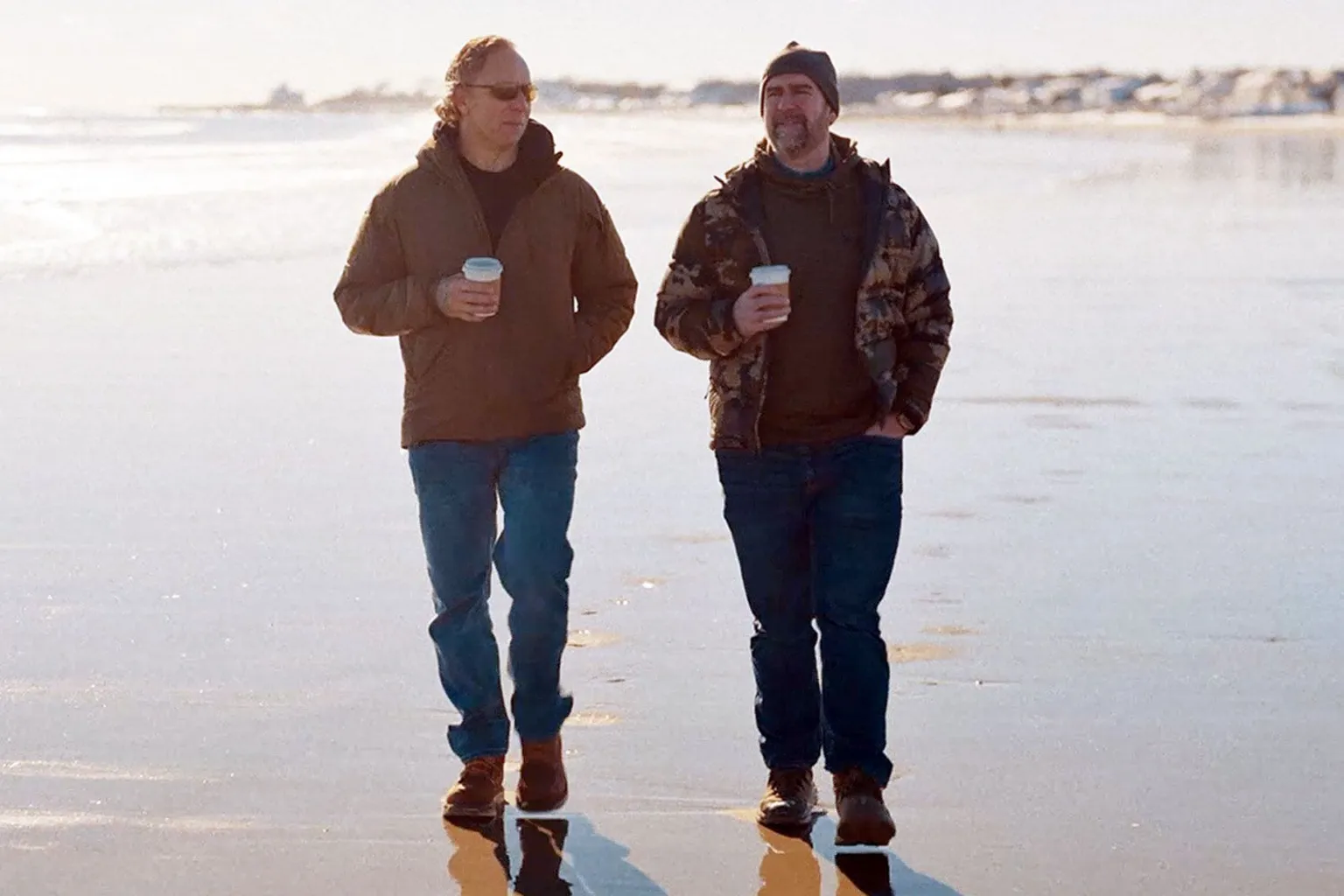 Greg Squire and Pete Manning walking along a beach holding takeaway drinks. They are both wearing blue trousers and brown jackets.