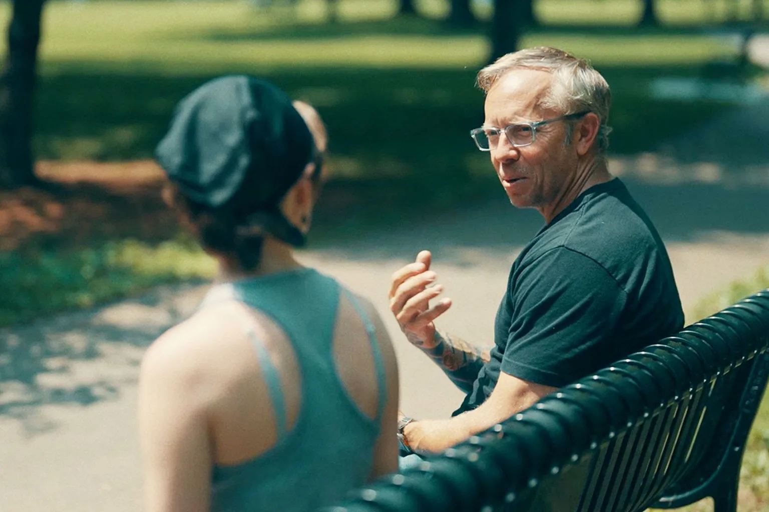 Squire and Lucy on a park bench talking. Squire is wearing a dark t-shirt and Lucy is wearing a cap, and blue racer-back top. 