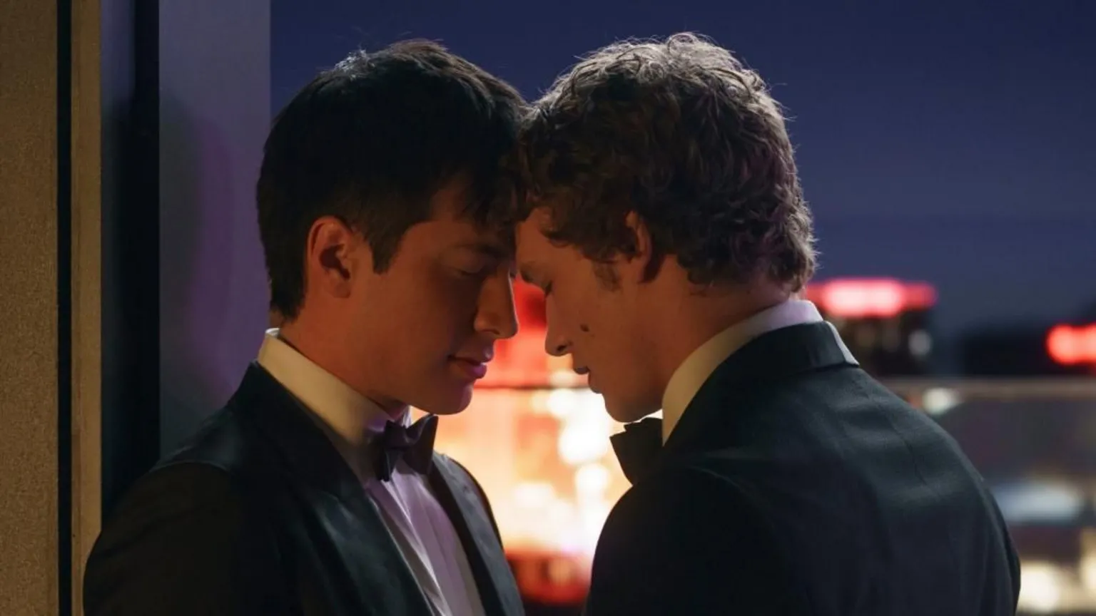 Two men wearing tuxedos with their heads touching on a balcony with the night sky and lit buildings behind them.