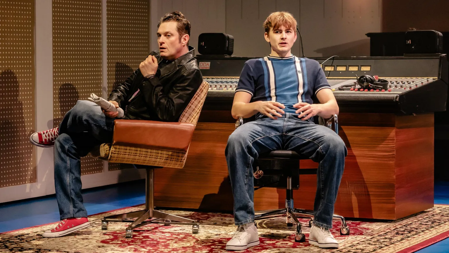 Danny With A Camera Matthew Horne in a leather jacket, jeans and red canvas trainers, next to Oscar Lloyd in striped blue T-shirt and jeans, both sitting in front of a recording mixing desk and speaking in a scene from The Battle
