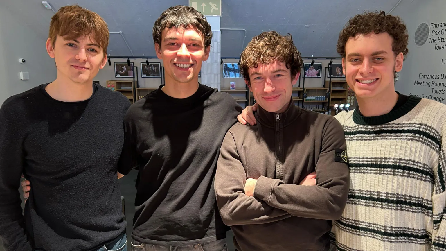 Four actors standing together in a row smiling with their arms around each other