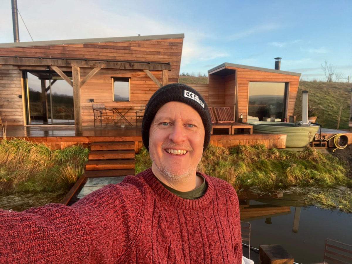 A man in a red jumper taking a selfie in front of a cabin