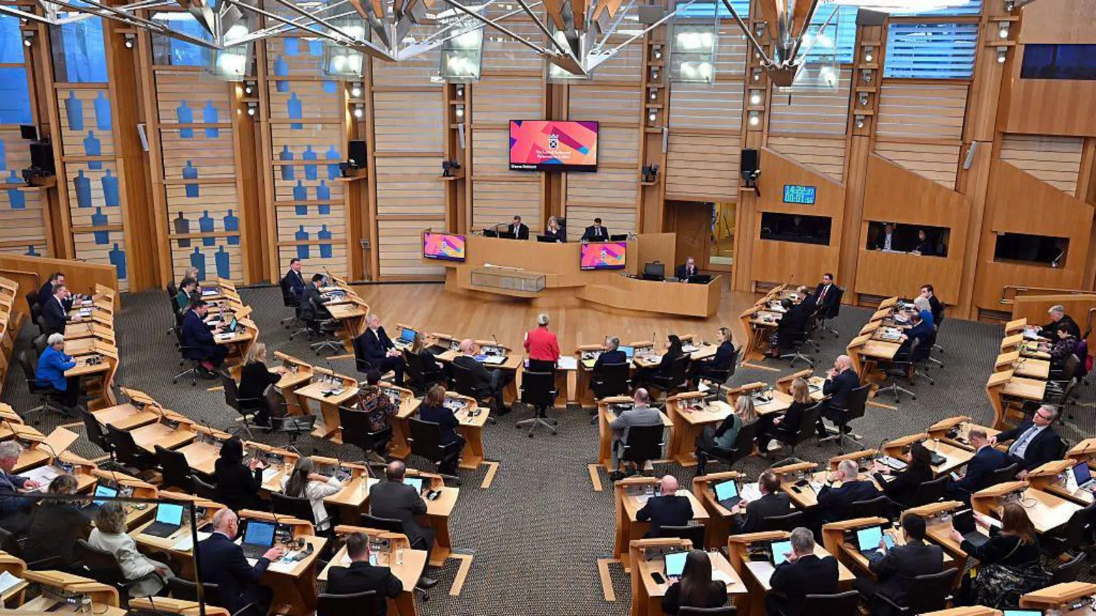 Finance Secretary Shona Robison (standing in the centre wearing a red jacket) delivers the Scottish Budget for 2026-2027 in the chamber of the Scottish Parliament in Edinburgh on 13 January, 2026. A number of MSPs can be seen at wooden desks with computer screens.