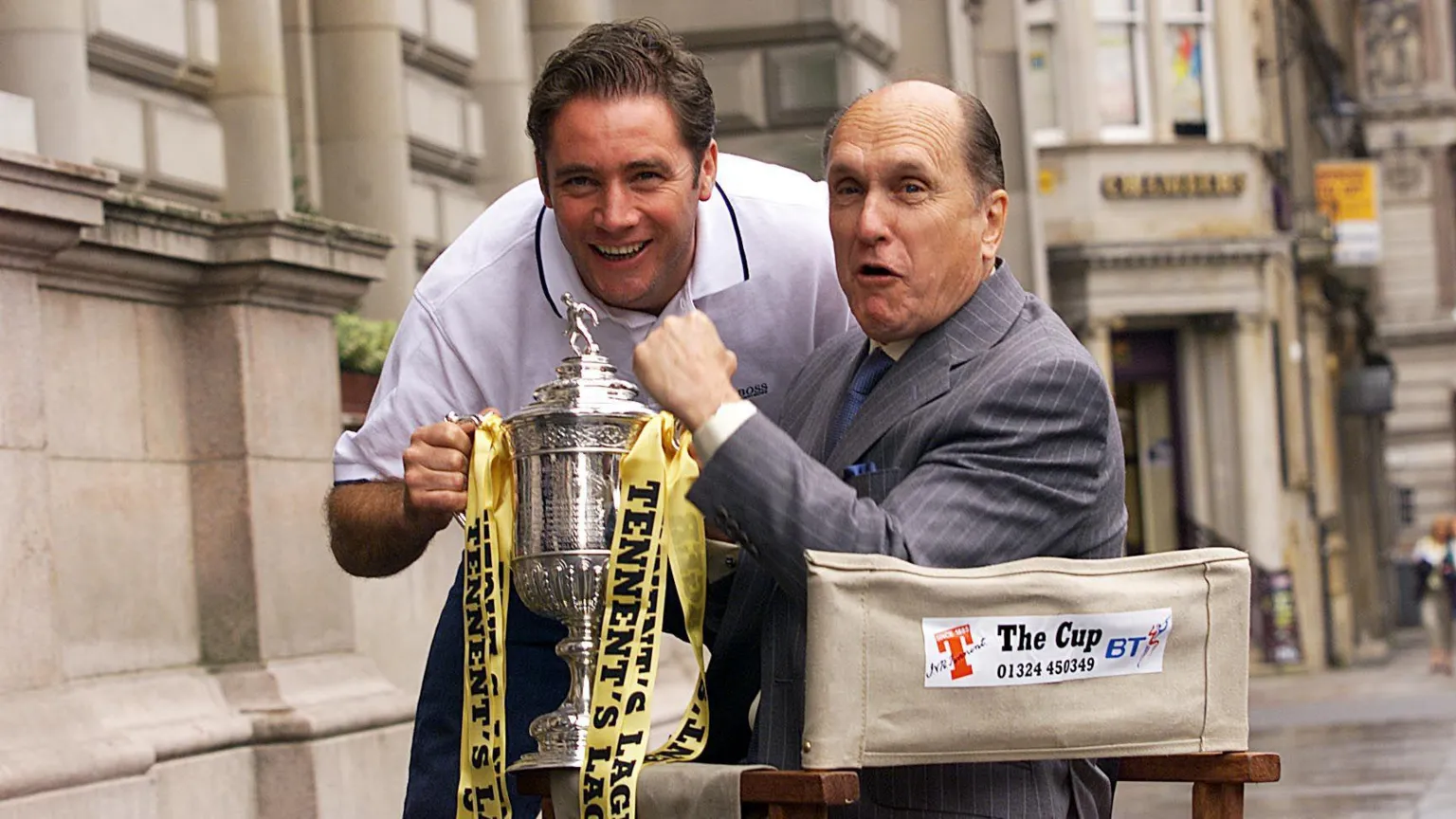 SNS Ally McCoist and Robert Duvall hold the Scottish Cup. Duvall is in a director's chair while McCoist is leaning forward in a white T-shirt.