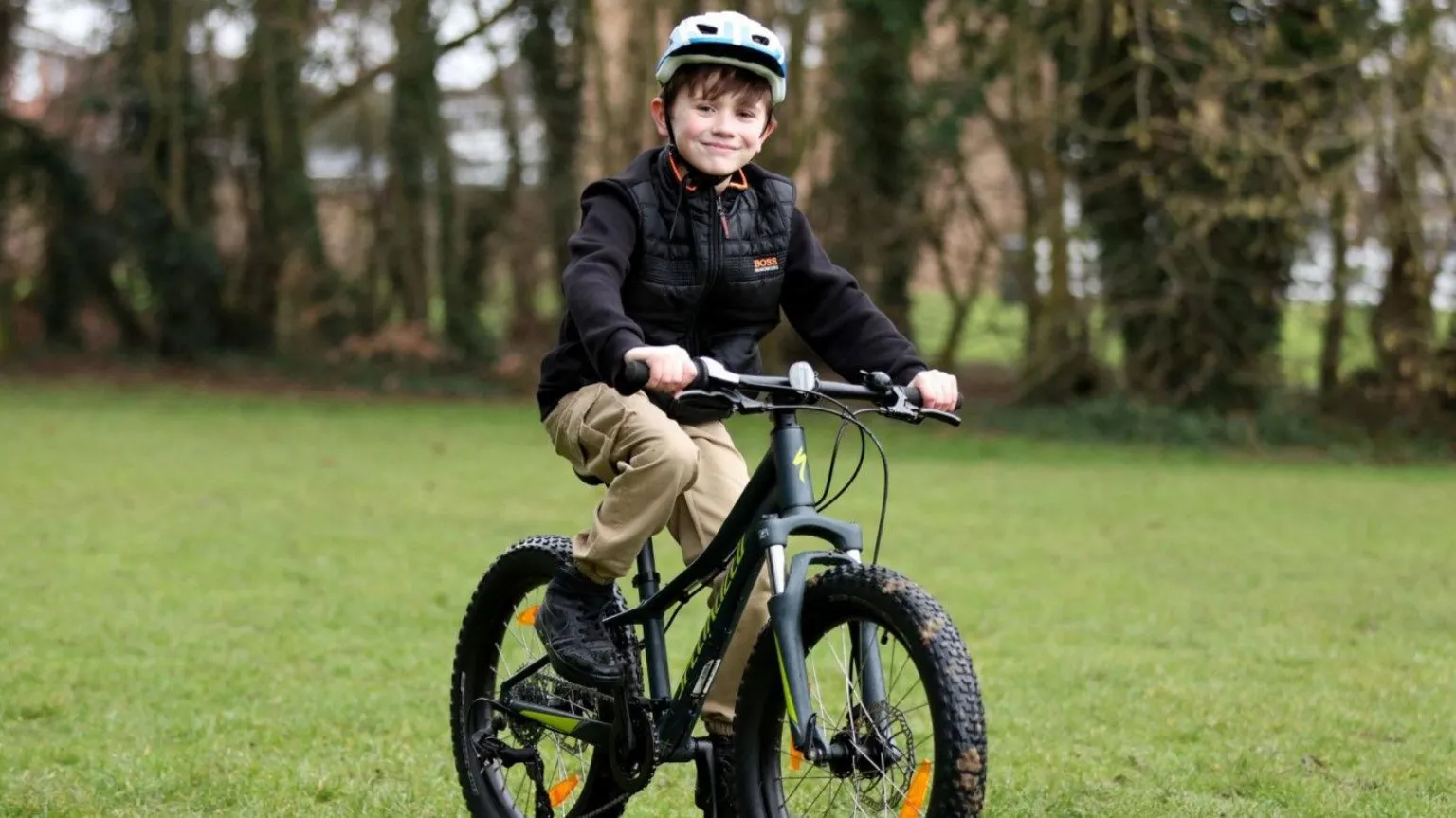 Chris Radburn/PA Wire Alfie is wearing a black jacket, beige trousers, black trainers and a cycling helmet. He is riding a bike on a field and is smiling at the camera.
