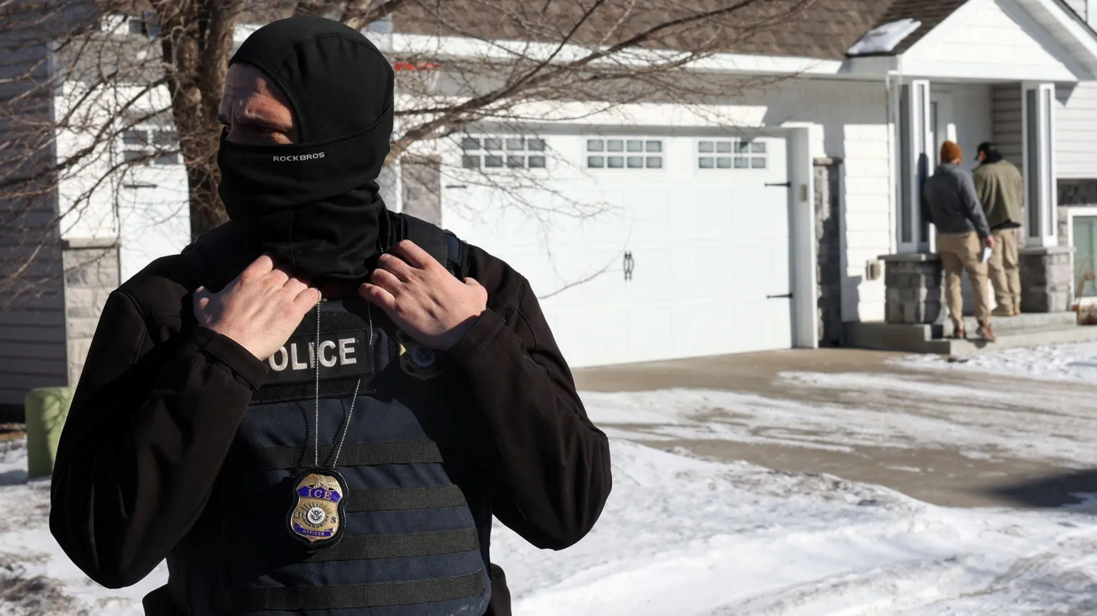  An ICE agent standing outside a white house. The agent has his face covered. 