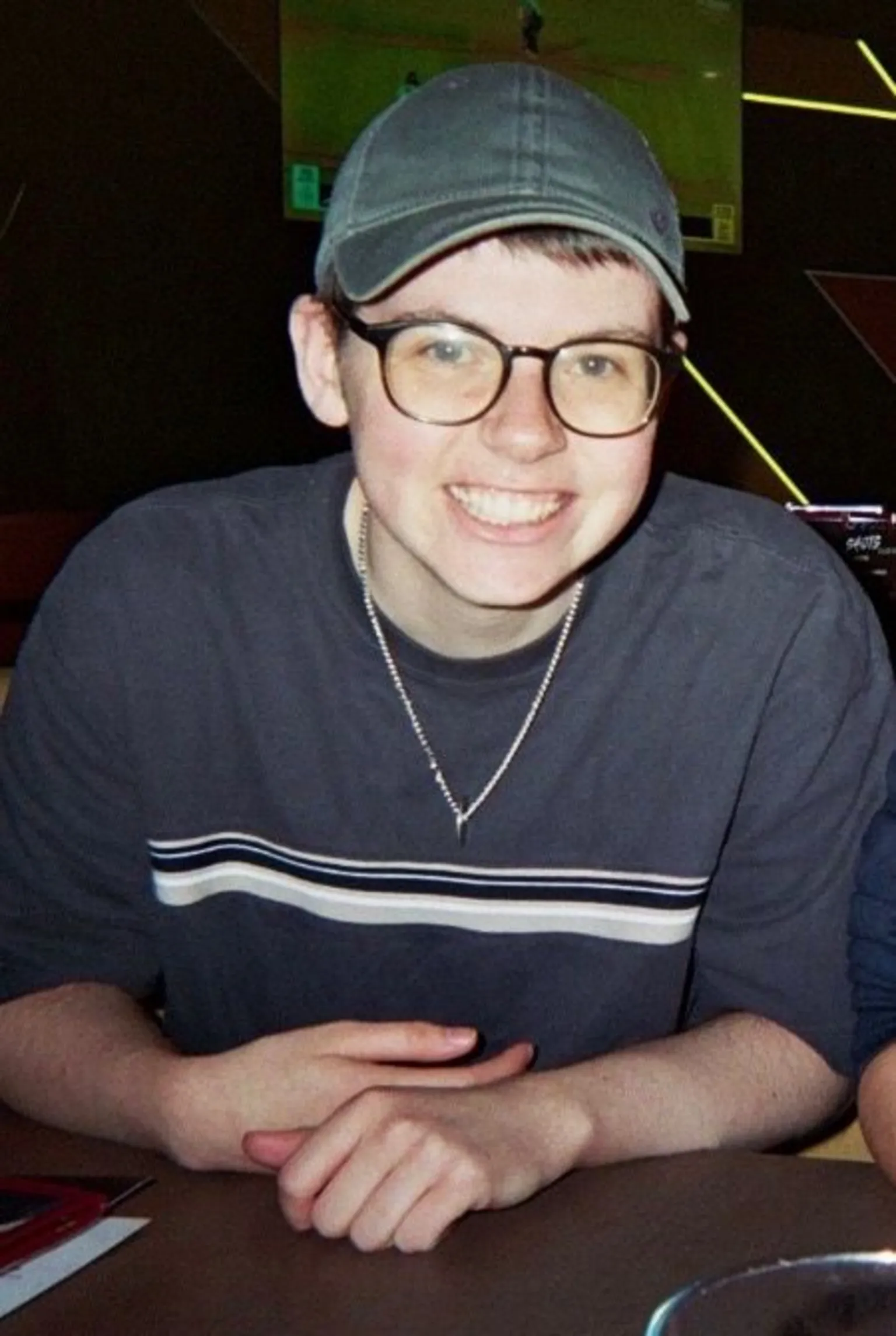 Alex Alex, a 21-year-old man, smiling at the camera. He is wearing a grey baseball cap and a grey t-shirt. He has glasses.