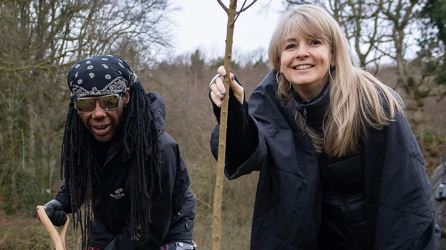 The Woodland Trust Nile Rodgers and Nancy Hunt plant a tree in woodlands near Derry. Nancy, with blonde hair and wearing a black coat is holding a spaling. Rodgers is on the left and wearing dark glasses , a head scarf and cblack jacket, and is holding a spade.