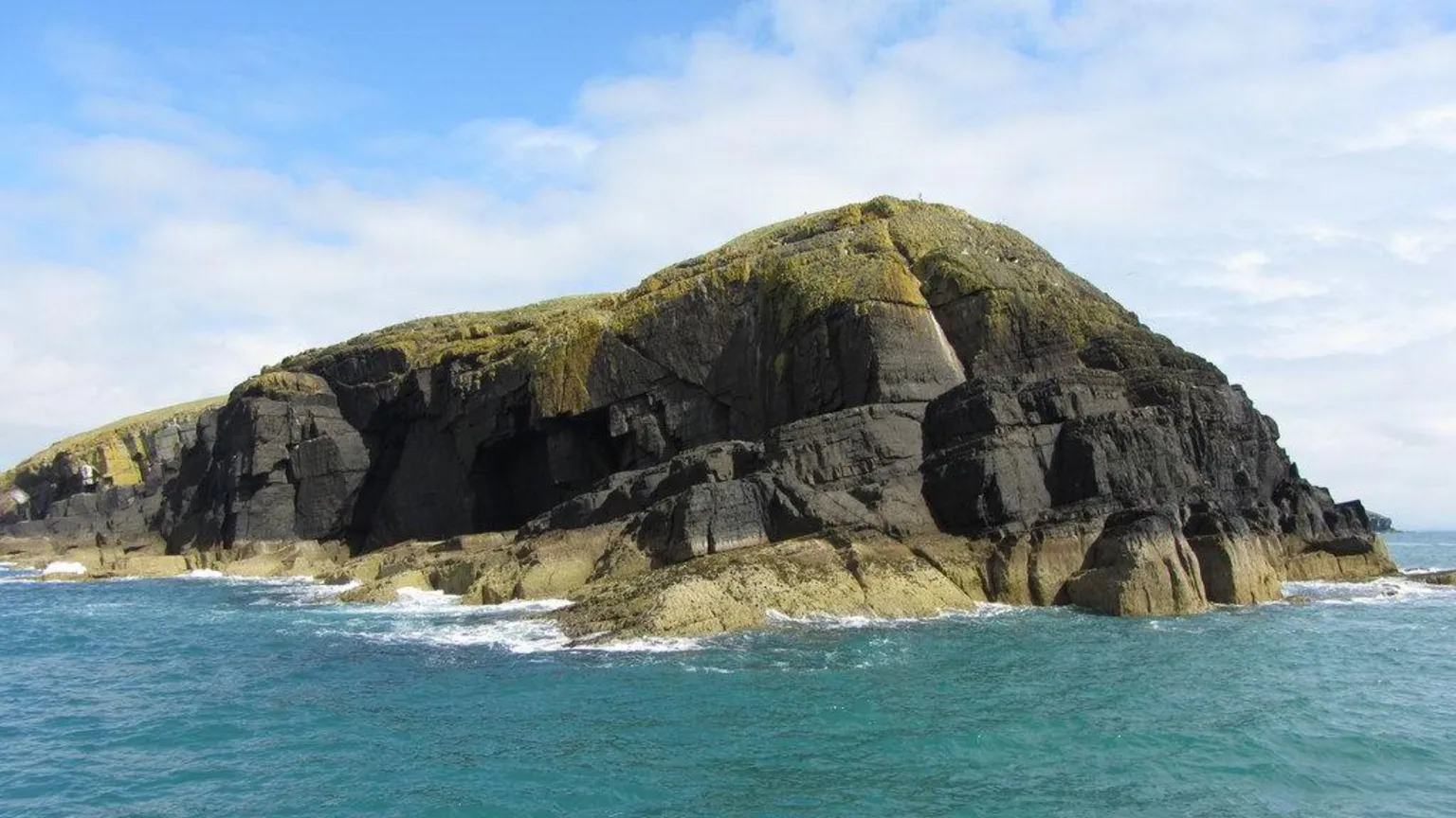 Geograph/Gareth James A view looking at the island from the sea. The rocky island rises up to a high cliff with rocks also jutting out into the sea, and greenery on top. There is a blue sky with white clouds, and the sea is turquoise. 