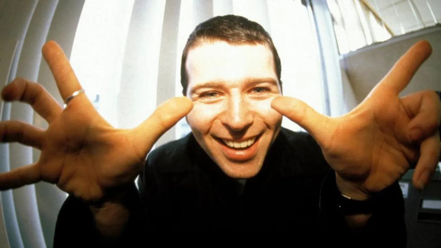 Mick Hutson/ Shows a man smiling with his fingers pointing upwards. He is wearing a black top.