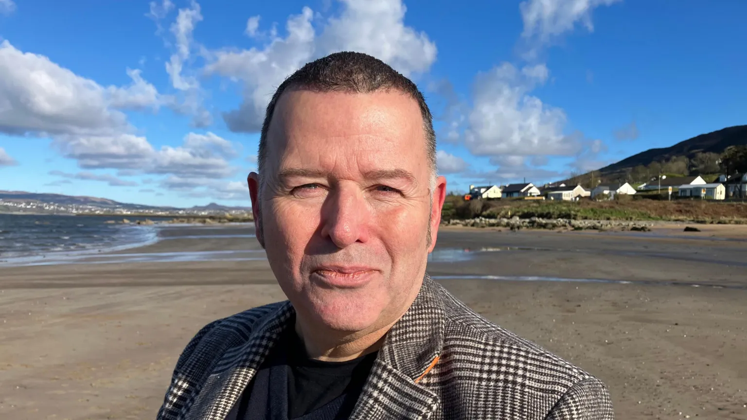 Peter Cunnah is shown on a beach with cloudy blue skies and a few houses in the background. 
