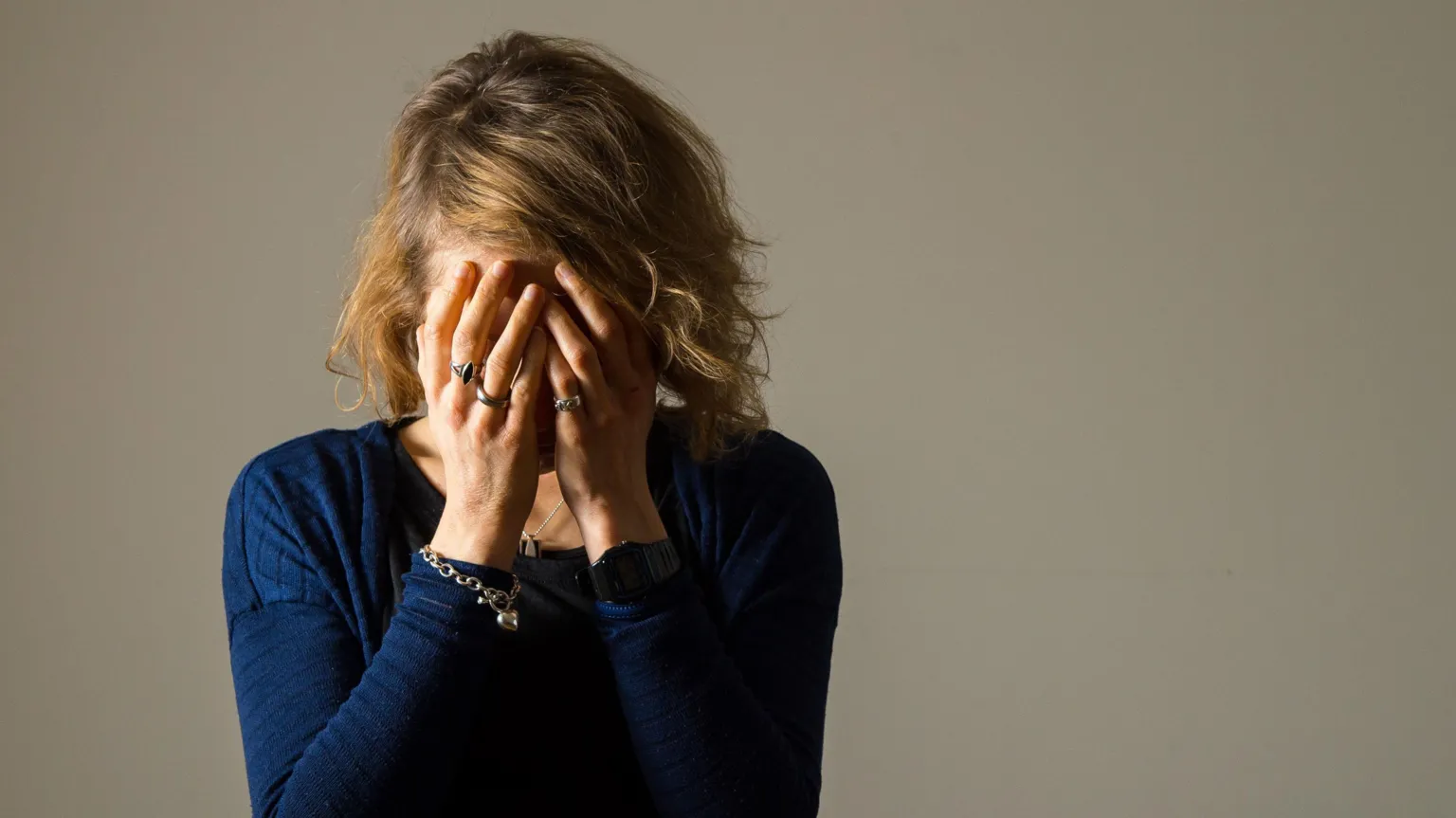 A woman with mid-length blonde hair is standing with her head in her hands. She is wearing a dark blue long-sleeved top. On her right wrist is a chunky silver bracelet made of links. She is also wearing a number of silver rings, one of which has a blue stone in it.