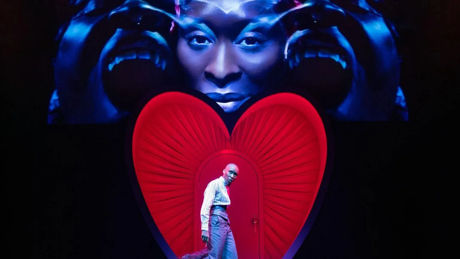 Daniel Boud Cynthia Erivo stands in front of a heart on stage in a suit while a visual projection of her face is above it. 