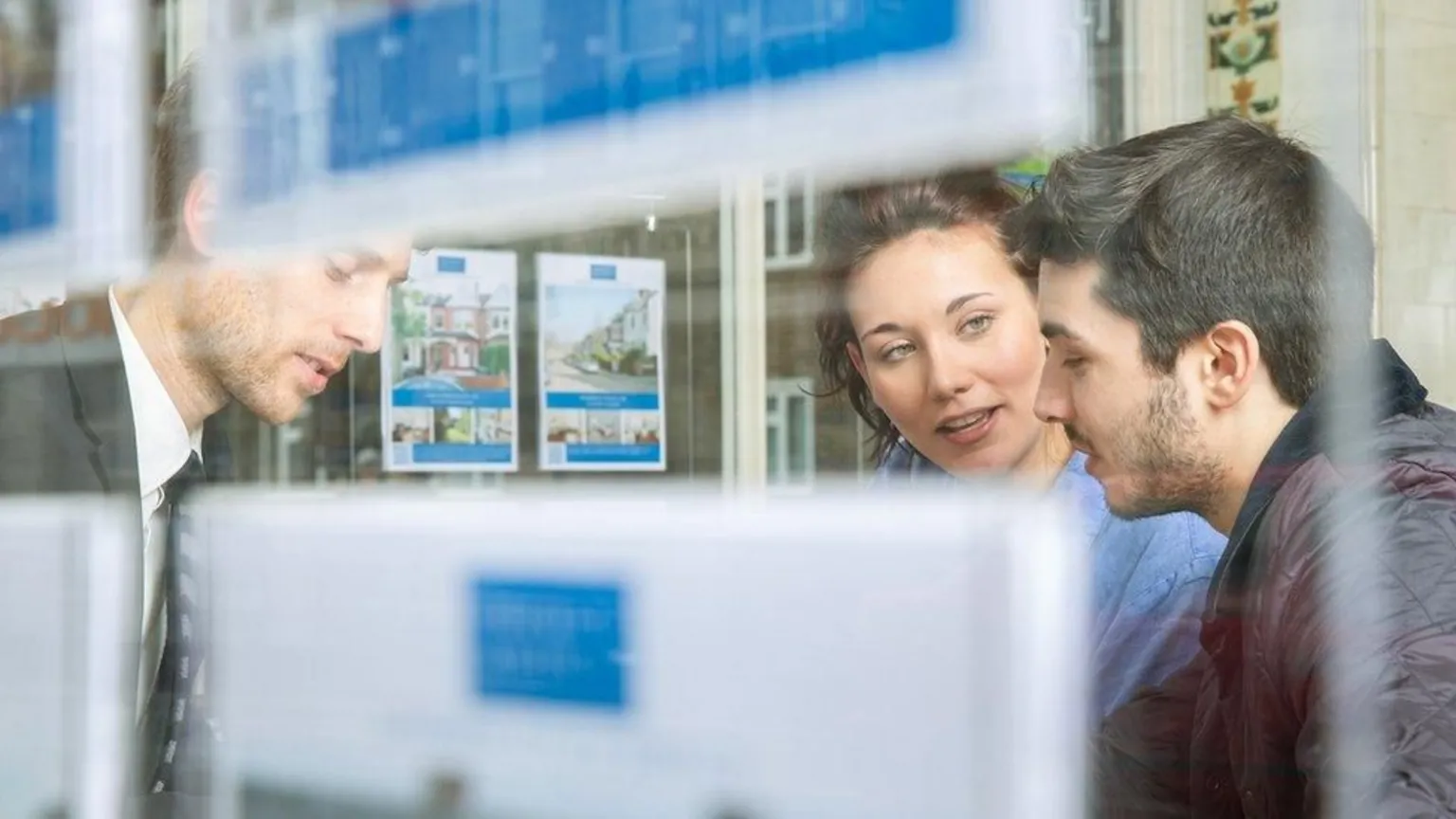  A picture looking through an estate agent's window showing a young couple talking to an estate agent who is wearing a grey suit