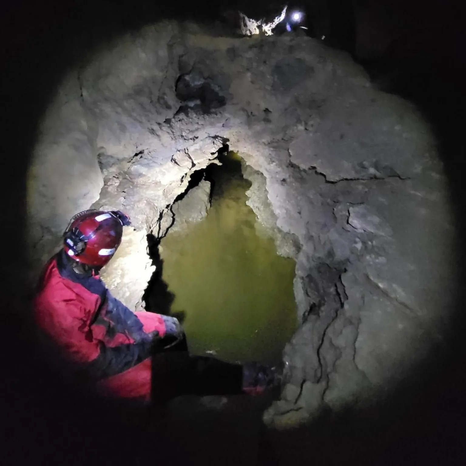 Assynt Mountain Rescue Team Torchlight illuminates a rescue team member and cave entrance with green water in front of it.