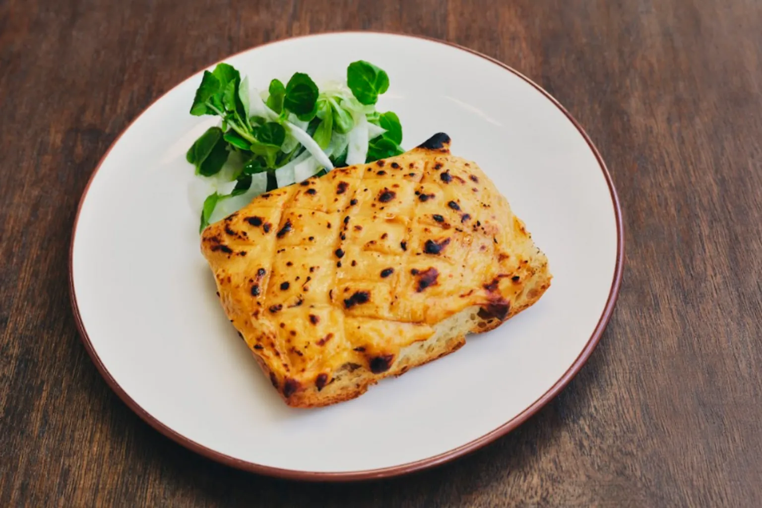 Michael Hani A round white plate sits on top of a brown, wooden, surface. On the plate is a yellow square of bread with melted cheese on top. To the side of the plate is a green leaf salad. 