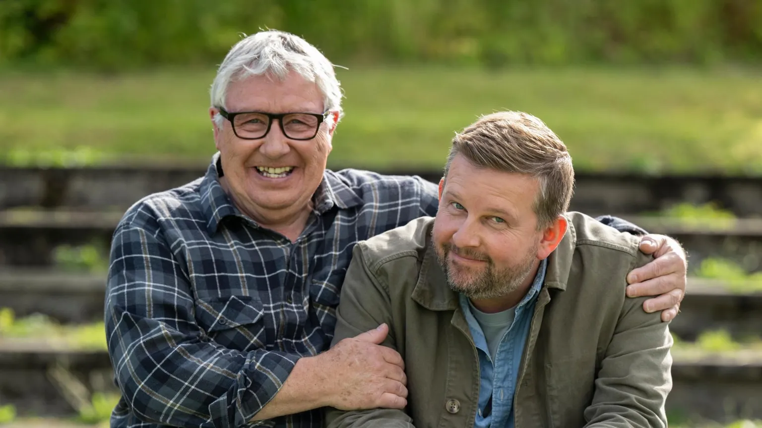Graeme Hunter / Happy Tramp North Gregor Fisher and Greg McHugh smiling at the camera. Fisher has his arm around McHugh. He wears a blue and white plaid shirt and glasses, while McHigh is wearing a green jacket over the top of a blue shirt.