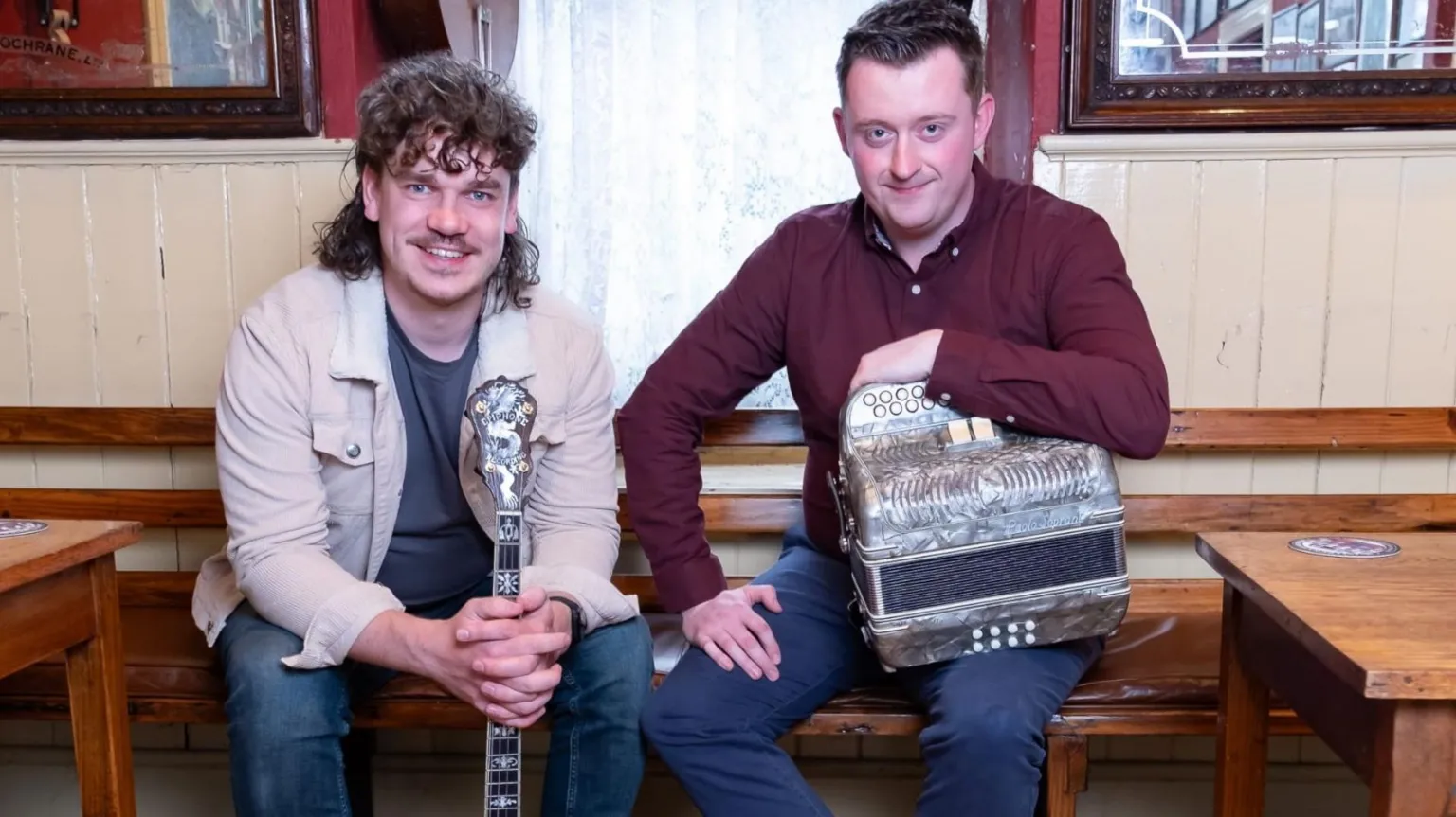 Photo by Ciaran Kelly Stephen is sitting on a wooden bench in a pub with his bandmate Gary Curley. Stephen, on the left, is wearing blue jeans. a grey t-shirt and a long-sleeved cream-coloured over-shirt. He is leaning over on his knees and holding a banjo upright between his legs. Gary has short brown hair, worn back off his face. He is holding his accordian between his left knee and left arm and wearing a maroon shirt and blue jeans. 
