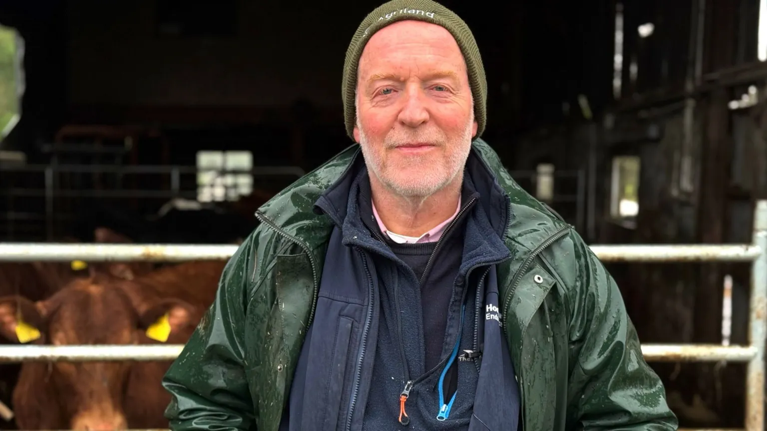 Richard Halleron is an older man. He is standing in front of a shed with metal gate which is closed in front of some brown cows. He has a short white beard and is wearing a green beanie hat, along with a dark green rain coat over a navy blue zip up top. 