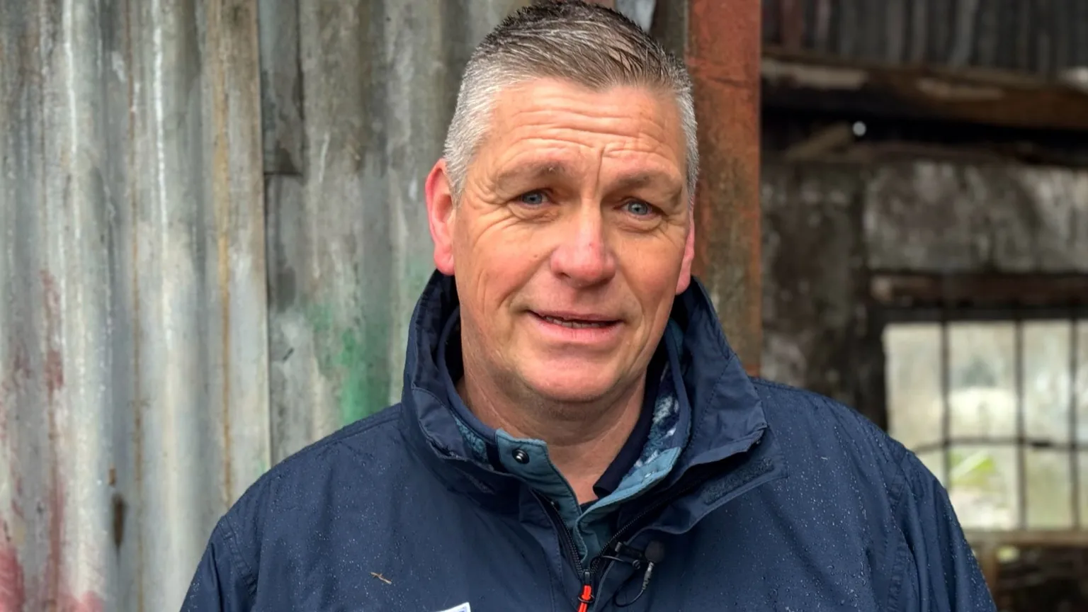 Dave Lowe has short brown hair on top swept over to the right, the hair on the sides of his head is grey. He is wearing a navy coat zipped up to the upturned collar. He is standing in front of a shed made out of corrugated metal. 