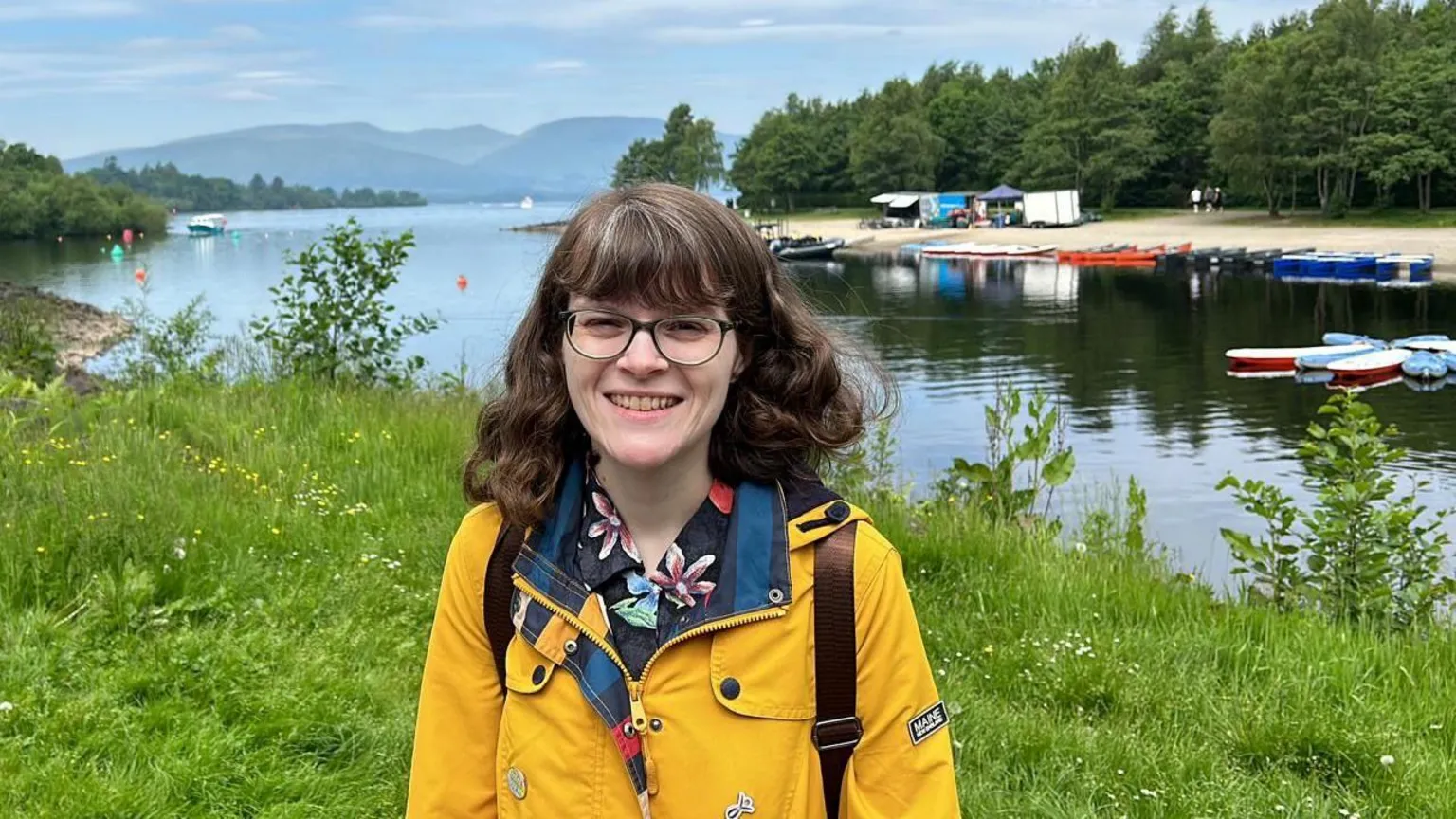 Louise Wallace A woman standing on the banks of a river, smiling. She has brunette hair to her shoulders, a yellow jacket and is wearing glasses.