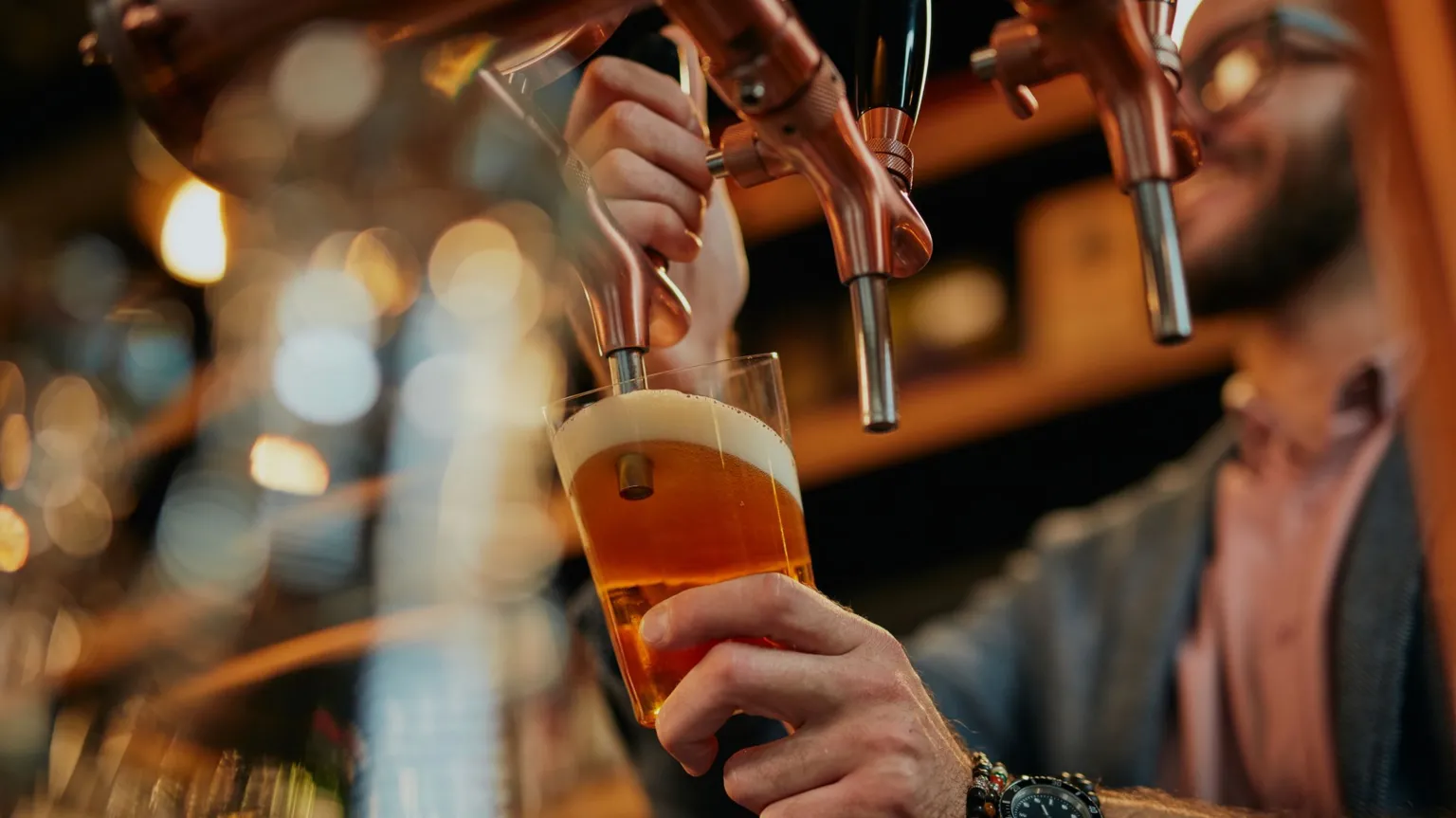  A man with a black beard and square glasses pulls a pint of lager. The glass is tilted underneath the pump and the beer has a foamy, white head. There are two other unused beer taps.