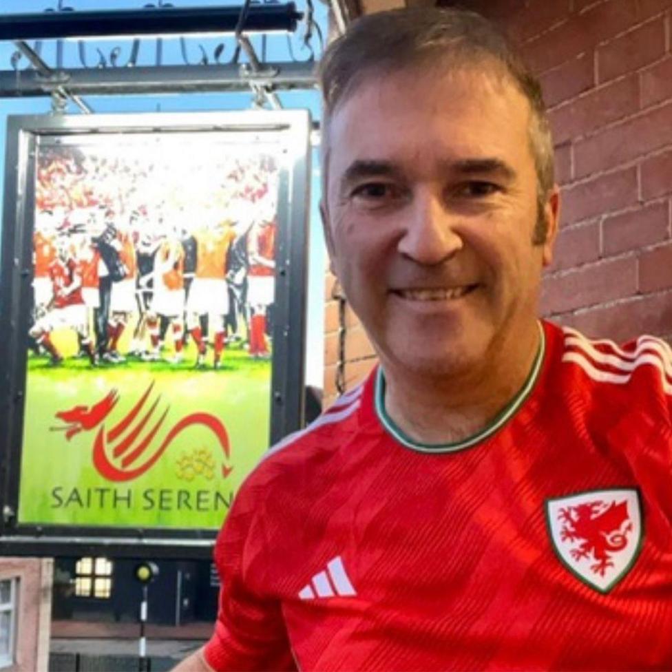 Chris Evans yn sefyll tu allan i'r Saith Seren, Wrecsam, yn gwisgo crys pêl droed Cymru