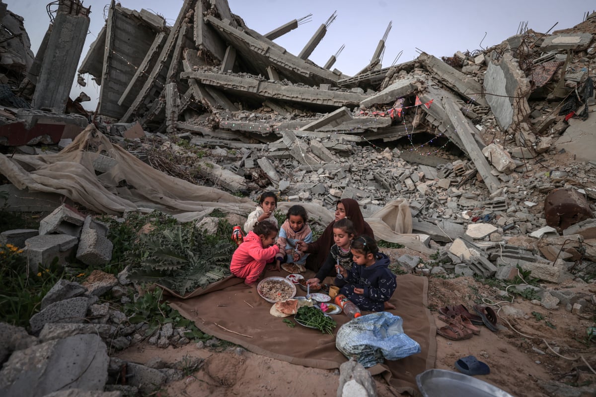 A Palestinian family of six, including young children, eat outside near the rubble of a wrecked building.