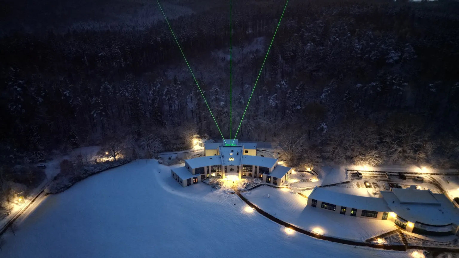 Gerd Baumgarten Three bright green lasers beam into the sky above a snowy landscape with brightly-lit buildings and forest behind them.