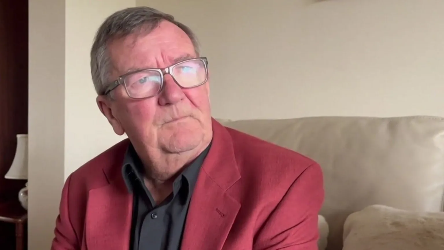 Brendan sat on his sofa looking at the camera. He is wearing a black shirt, grey glasses and a maroon suit jacket. He has short grey hair. 