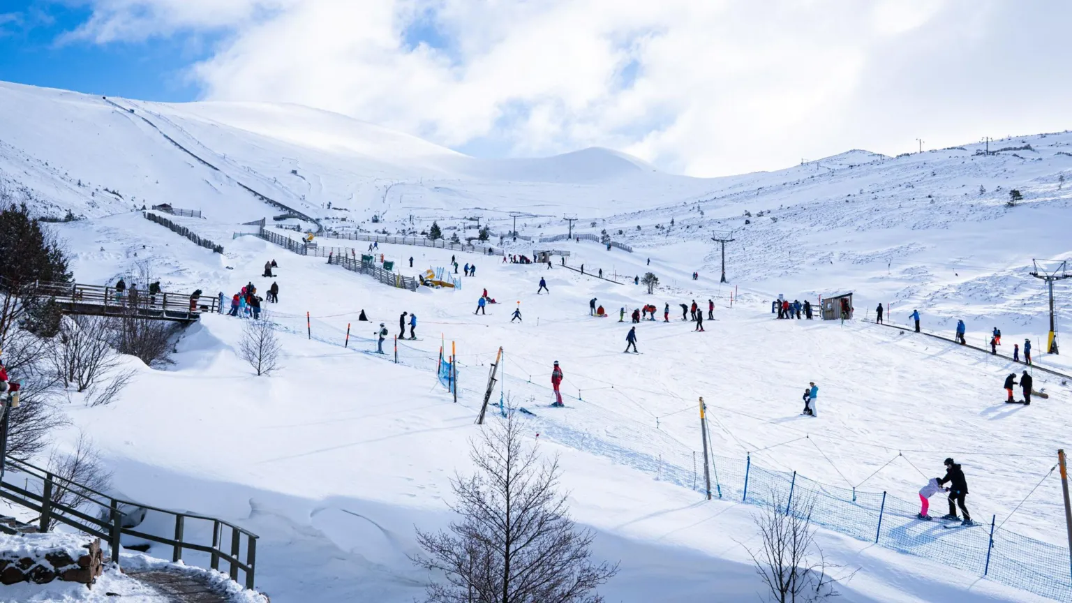 Angus Trinder/Cairngorm Mountain Snowsports enthusiasts at Cairngorm Mountain. The skiers and snowboarders are dotted across snow-covered slopes. It is a bright, sunny day.