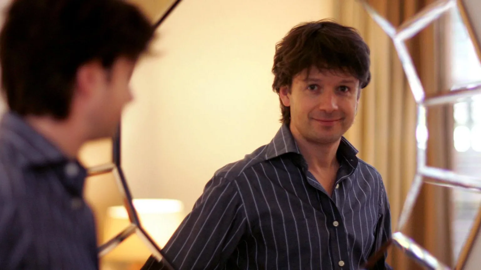 City AM/Shutterstock A man in a blue pinstriped shirt smiles in a reflection in the mirror.