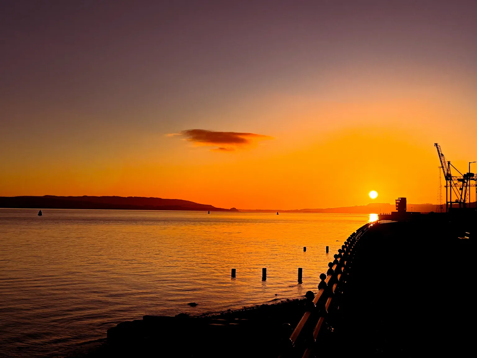 Tommy Bamford Sun rising over the Firth of Clyde. The sky is orange and is reflecting on the water. In the distance there are cranes in silhouette .