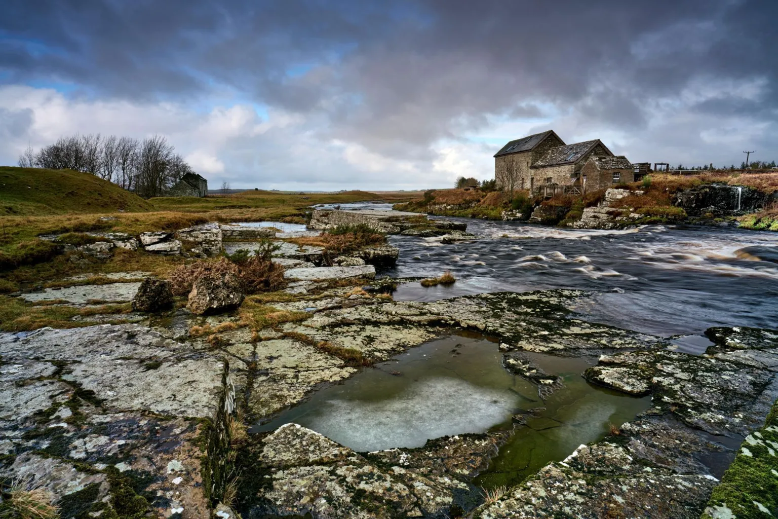 John Cuthbert The stone mill building at Hawick can be seen across a river, with frozen puddles of water between the rocks in the foreground. 