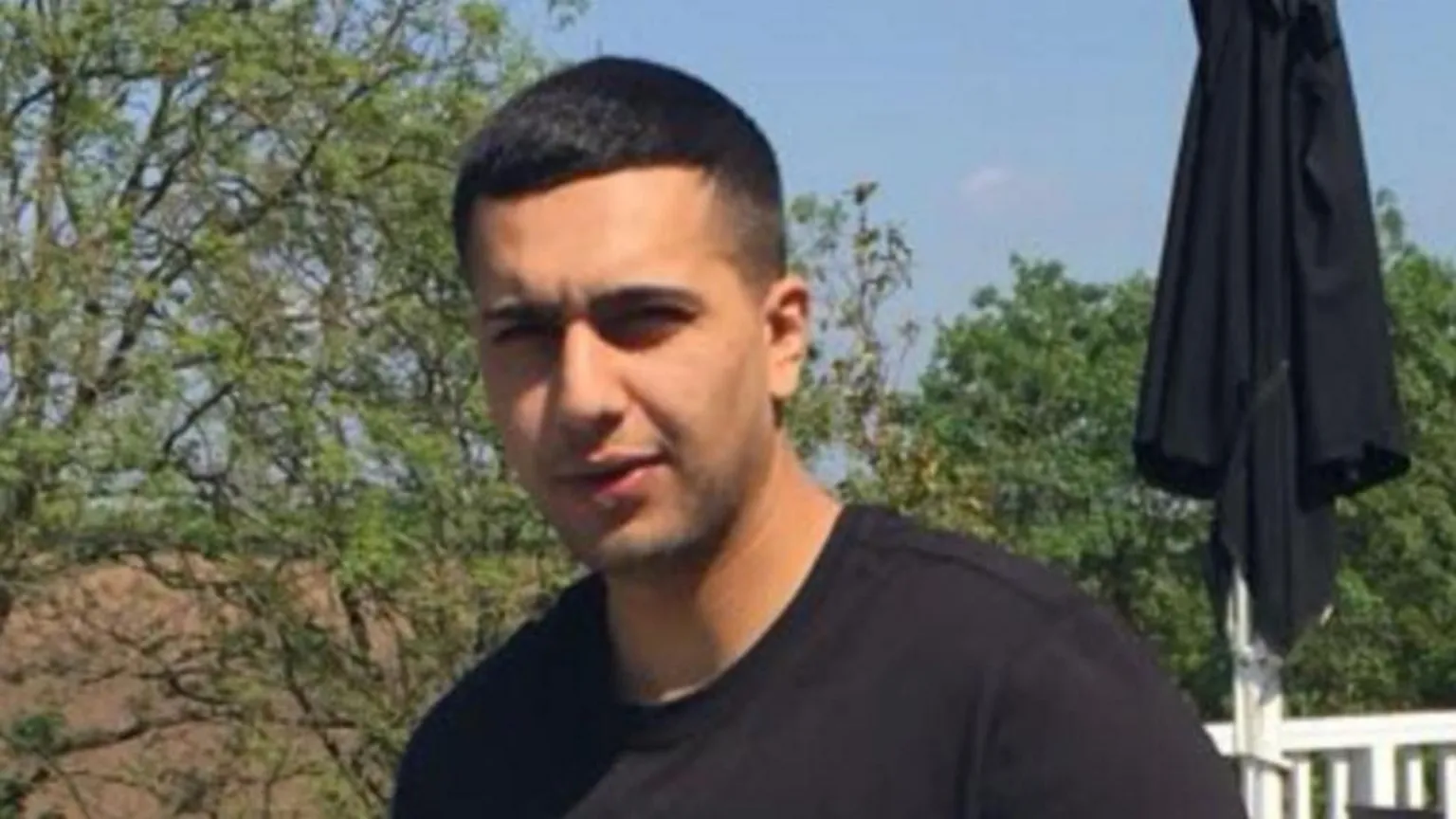 South Wales Police Image of Kamran Aman wearing a black t shirt and has short cropped hair. There are trees and a white railing behind him. 