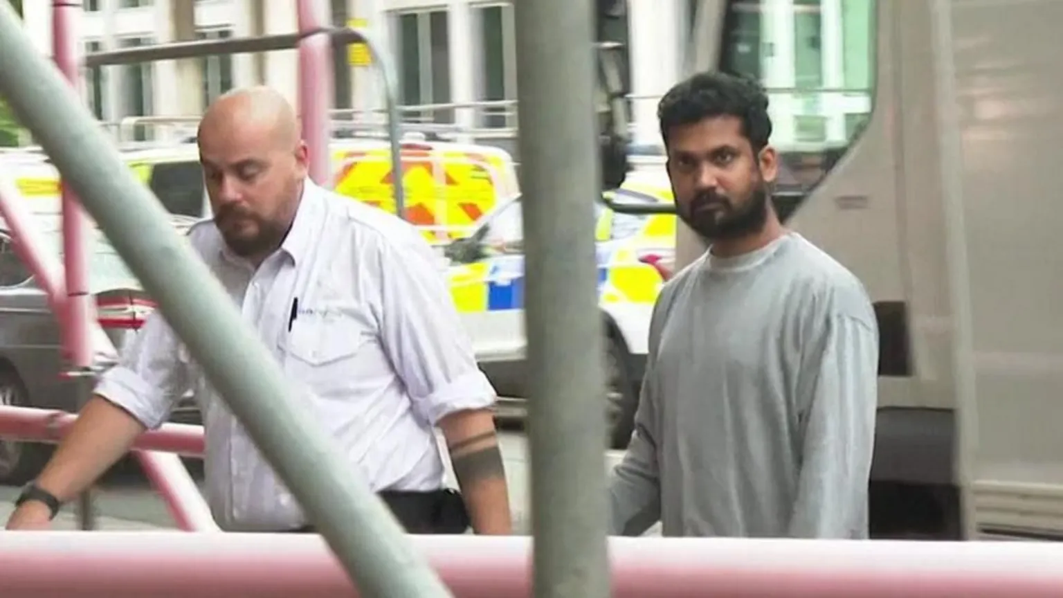 Weragalage handcuffed to an officer walking from a police van into Cardiff Crown Court. He is wearing grey prison uniform and is looking at the camera. He has short dark brown hair and a beard. 