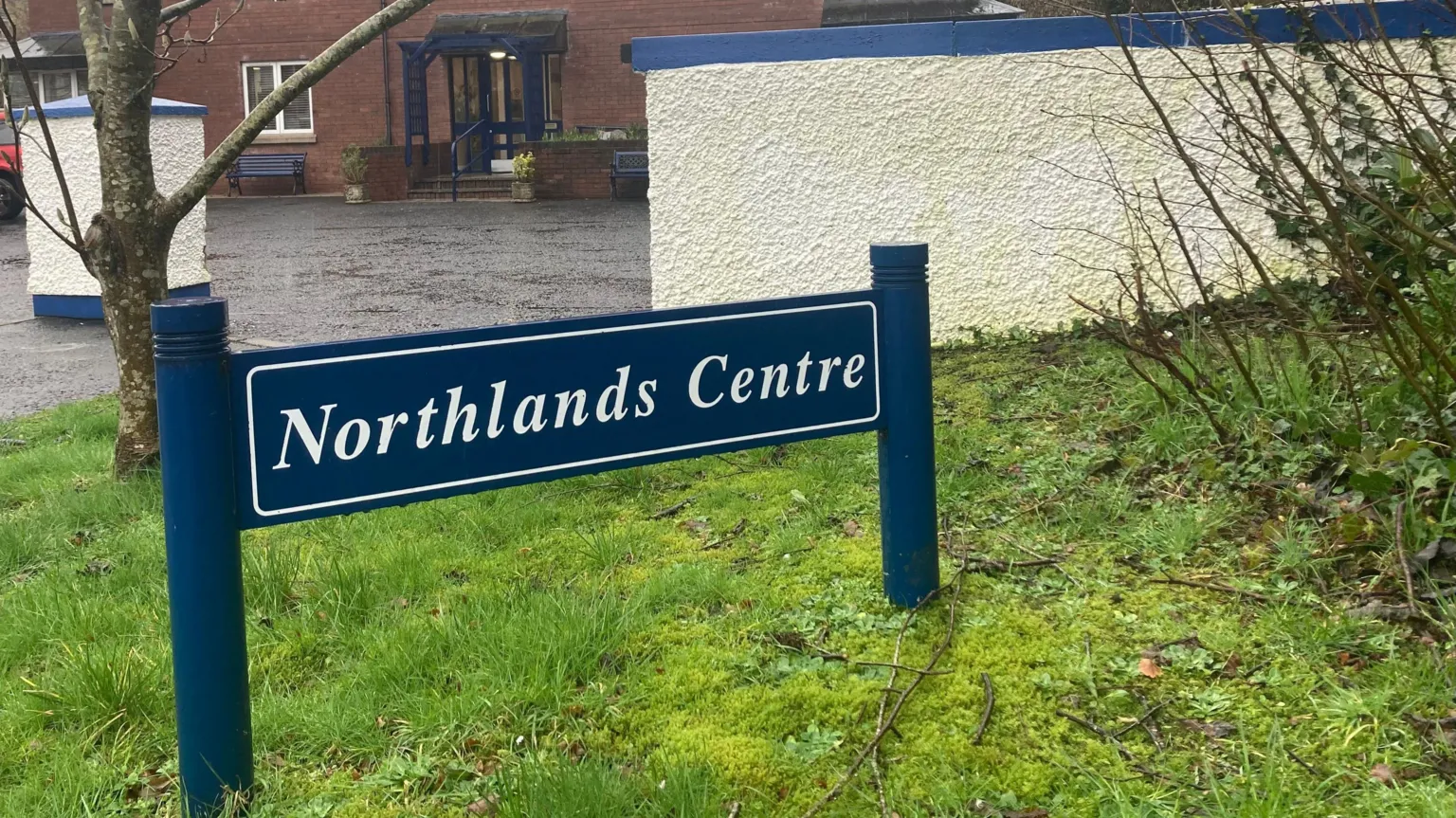 A dark blue sign states Northlands Centre in white lettering. There is a building in the background. The sign has been erected on grass.