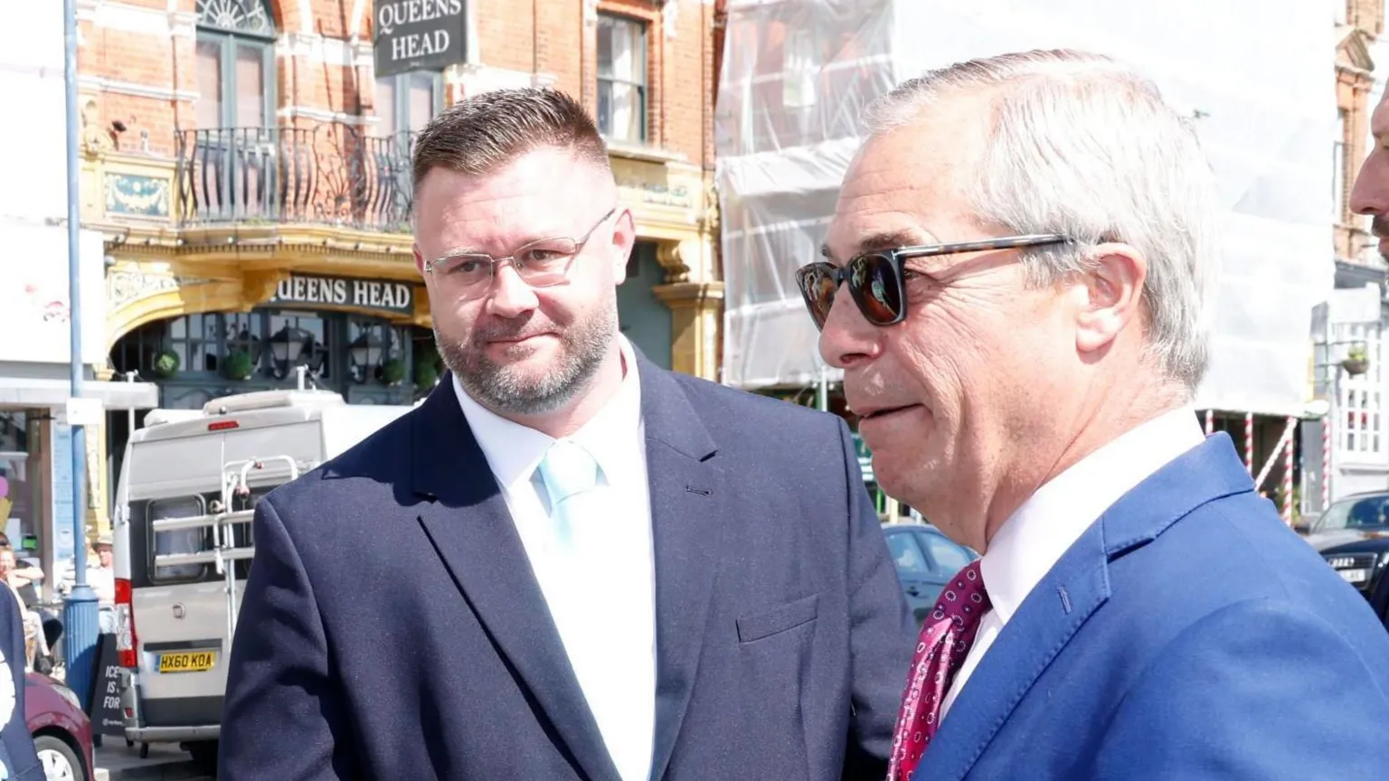 SteveFinnPhotography Taylor, wearing a suit with a light blue tie, with a street seen in the background. He is stood next to Nigel Farage, who is wearing a suit and sunglasses.