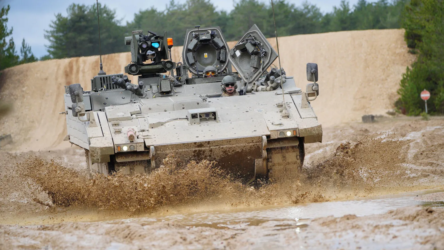  Soldiers in helmets and goggles riding in a khaki Ajax armoured vehicle. Mud is flicking up from the wet ground around the vehicle as it is in motion.