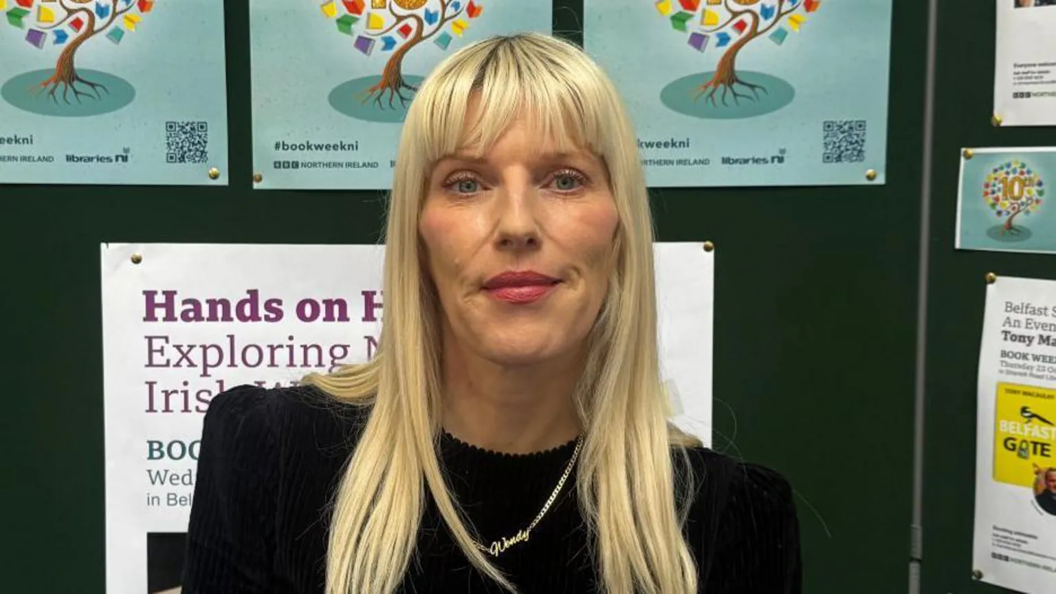 Wendy Erskine has long blonde hair. She is wearing a black dress and is smiling at the camera. There are posters for Book Week NI in the background. 